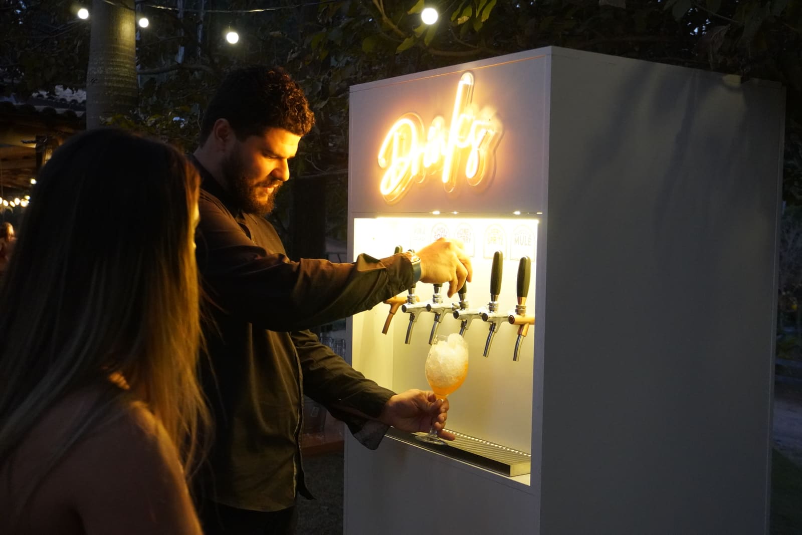 Convidado se servindo de um drink no totem iluminado durante uma festa ao ar livre, à noite
